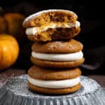 A stack of three vegan pumpkin whoopie pies on a metal plate, one with a bite taken out of it.