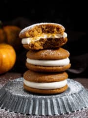 A stack of three vegan pumpkin whoopie pies on a metal plate, one with a bite taken out of it.