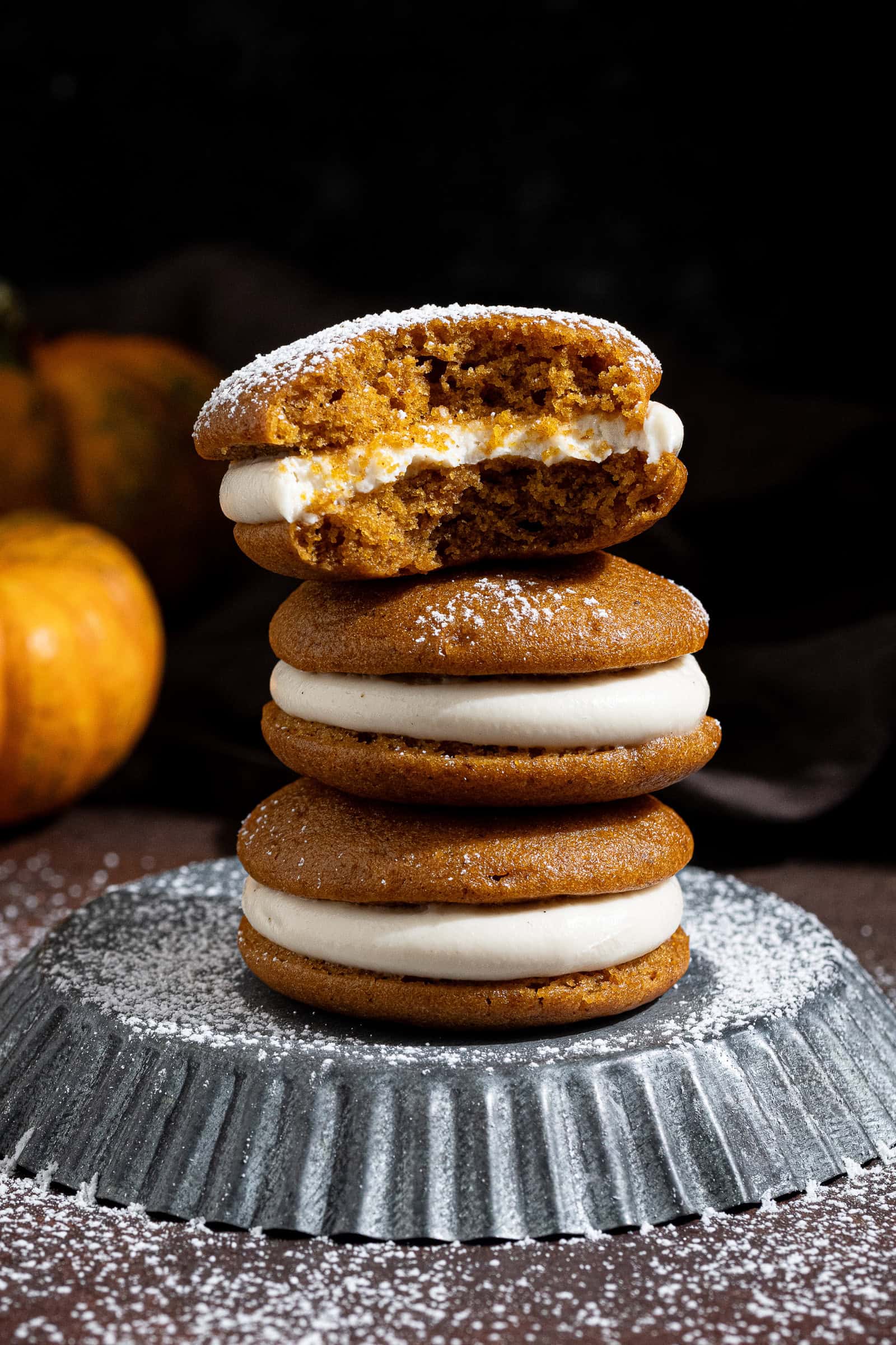 A stack of three vegan pumpkin whoopie pies on a metal plate, one with a bite taken out of it.