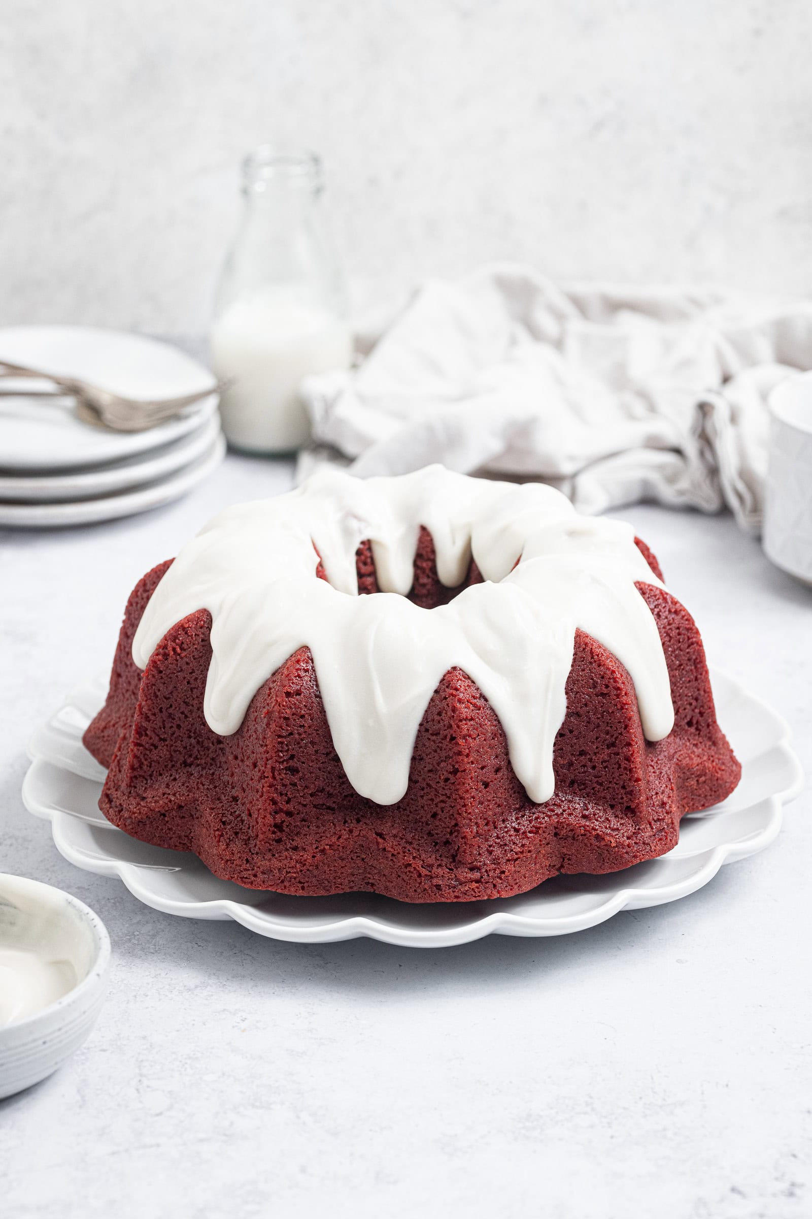 Vegan red velvet bundt cake on a white plate on a white background with white plates and a glass milk bottle in the background.
